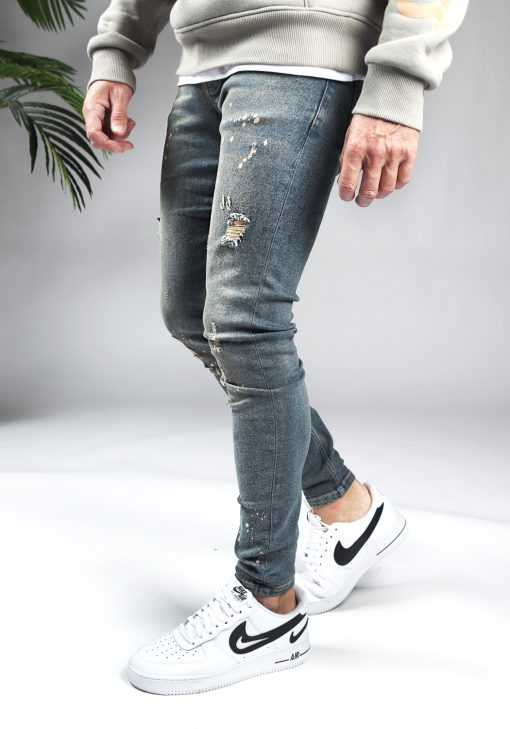 Rechter zijaanzicht damaged heren skinny jeans met zandkleurige details. Voorzien van vier zakken, knoopsluiting en riemlussen. Gecombineerd met crèmekleurige hoodie en witte sneakers.
