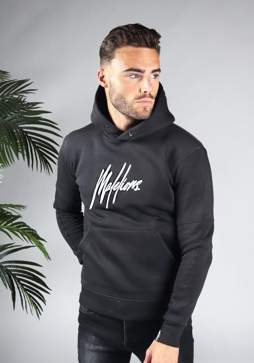 Vooraanzicht van model gekleed in zwarte hoodie met het grote witte Malelions logo op de borst. Het model heeft een arm achter zijn rug en kijkt naar links.