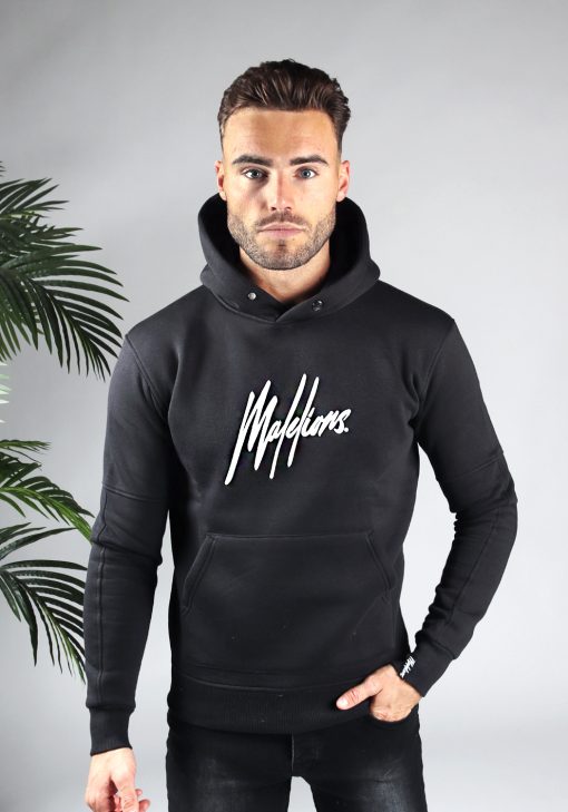 Vooraanzicht van model gekleed in zwarte hoodie met het witte Malelions logo op het midden van de borst in combinatie met zwarte jeans. Het model heeft een hand in zijn broekzak en kijkt recht in de camera.