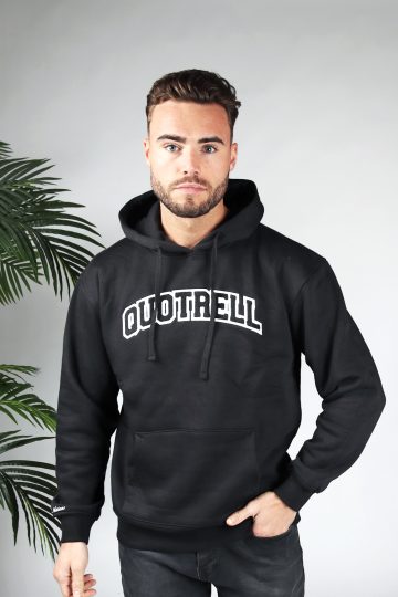 Vooraanzicht van model gekleed in een zwarte hoodie met daarop de tekst quotrell in university stijl. Het model heeft een hand in zijn broekzak en kijkt recht in de camera.