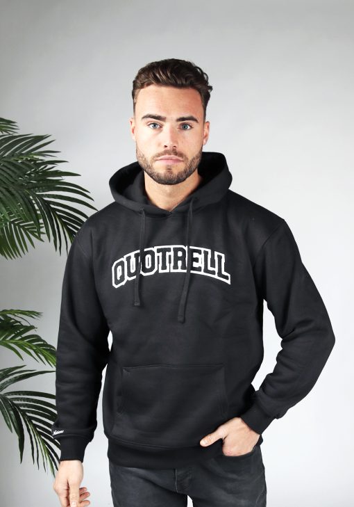 Vooraanzicht van model gekleed in een zwarte hoodie met daarop de tekst quotrell in university stijl. Het model heeft een hand in zijn broekzak en kijkt recht in de camera.