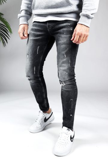 Linker zijaanzicht zwarte denim damaged heren skinny jeans, gemaakt van stretch stof. Voorzien van vier zakken, knoopsluiting en riemlussen. Gecombineerd met grijze hoodie en witte sneakers.