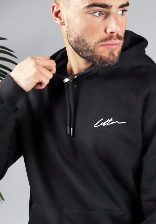 Close up van model gekleed in een zwarte hoodie met het signature cotton logo op de borst. Model heeft een hand bij de hals van de hoodie en kijkt naar links.