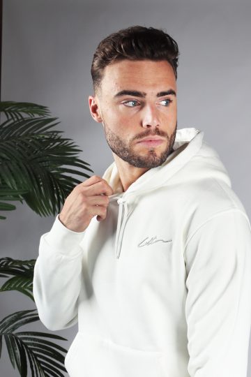 Close up van model gekleed in een witte hoodie met het witte signature cotton logo op de borst. Model heeft een hand bij de hals van de hoodie en kijkt naar links.