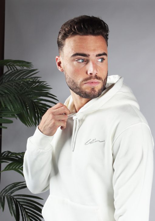 Close up van model gekleed in een witte hoodie met het witte signature cotton logo op de borst. Model heeft een hand bij de hals van de hoodie en kijkt naar links.