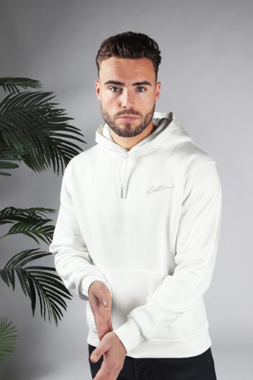 Vooraanzicht van model gekleed in een witte hoodie met het witte signature cotton logo op de borst en een zwarte broek. Model heeft zijn handen voor zich en kijkt in de camera.