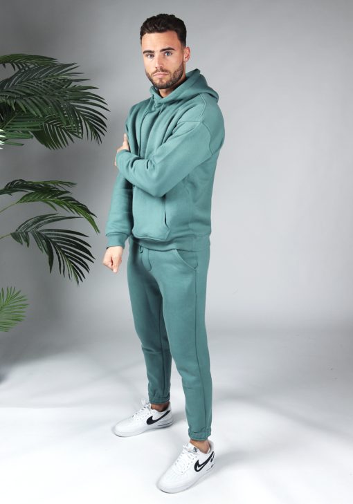 Schuin aanzicht full body shot van model gekleed in een turquoise fleece katoen tracksuit gecombineerd met witte sneakers. Het model houdt een hand over zijn bovenarm en kijkt recht in de camera.