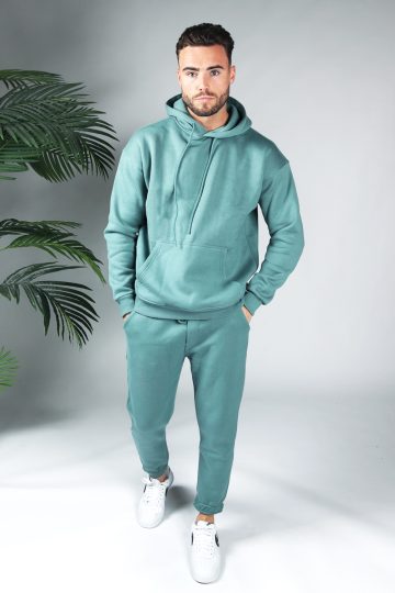 Full body shot van model gekleed in een turquoise fleece katoen tracksuit gecombineerd met witte sneakers. Het model heeft zijn handen in zijn zakken en kijkt recht in de camera.