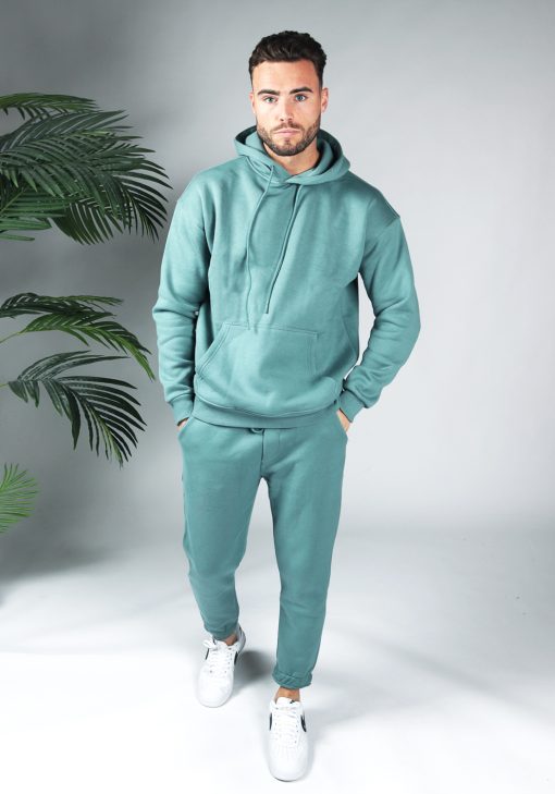 Full body shot van model gekleed in een turquoise fleece katoen tracksuit gecombineerd met witte sneakers. Het model heeft zijn handen in zijn zakken en kijkt recht in de camera.