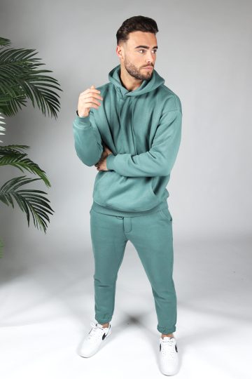 Full body shot van model gekleed in een turquoise fleece katoen tracksuit gecombineerd met witte sneakers. Het model heeft een arm rond zijn middel en kijkt naar links.
