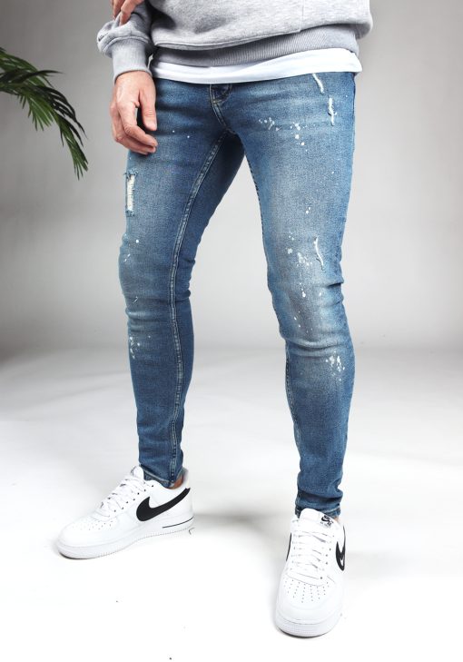 Voorkant denim heren skinny jeans met damaged look en verfspetters. Gemaakt van stretch stof en voorzien van vier zakken, knoopsluiting en riemlussen. Gecombineerd met grijze hoodie en witte sneakers.