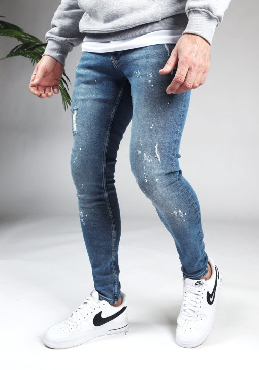 Linker zijaanzicht denim heren skinny jeans met damaged look en verfspetters. Gemaakt van stretch stof en voorzien van vier zakken, knoopsluiting en riemlussen. Gecombineerd met grijze hoodie en witte sneakers.
