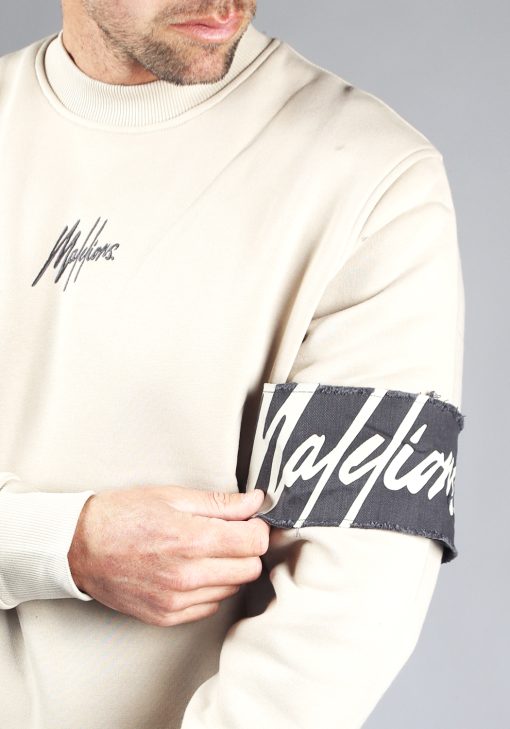 Close up van beige Malelions sweater gecombineerd met een zwarte pet. De sweater heeft een zwarte band met het witte logo om de linkerarm en het kleine zwarte logo op de borst. Het model pakt de zwarte band rond zijn arm vast.