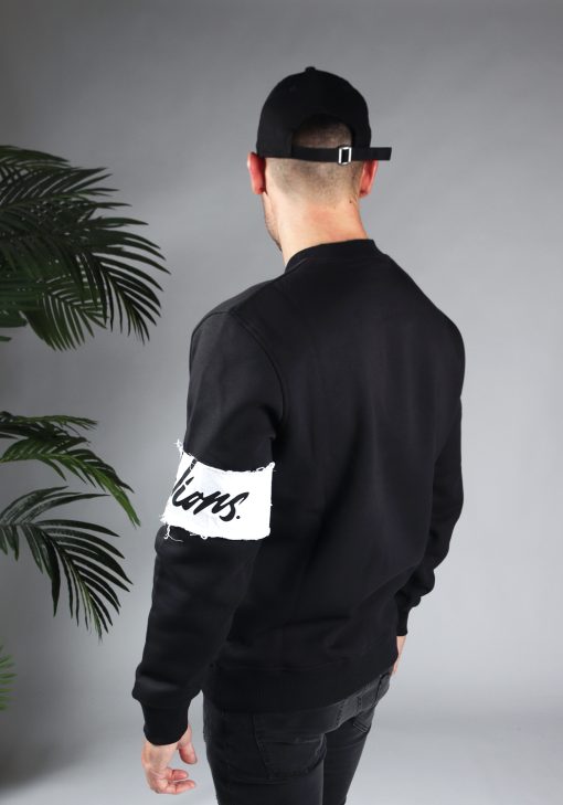 Achteraanzicht van model gekleed in de zwarte Malelions sweater in combinatie met zwarte jeans en witte sneakers. De trui heeft een witte band met het donkere logo om de linkerarm. Het model pakt zijn arm vast en kijkt naar de grond.