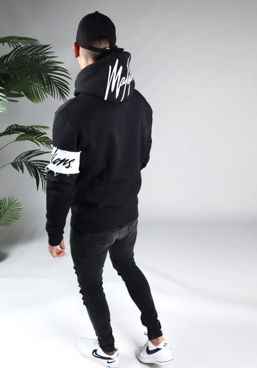 Achteraanzicht van model gekleed in de zwarte Malelions hoodie. De trui heeft een witte band met het donkere logo om de linkerarm en het grote logo op de capuchon. Het model heeft zijn armen langs zich en kijkt naar de grond.