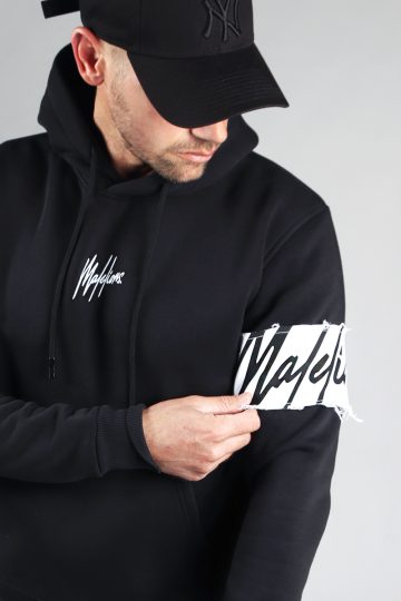 Close up van model gekleed in de zwarte Malelions hoodie. De trui heeft een witte band met het donkere logo om de linkerarm. Het model grijpt naar de witte band om zijn arm.