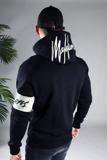 Achteraanzicht van model gekleed in de donkerblauwe Malelions hoodie. De trui heeft een witte band met het donkere logo om de linkerarm en het grote logo op de capuchon. Het model heeft zijn armen langs zich en kijkt naar de grond.