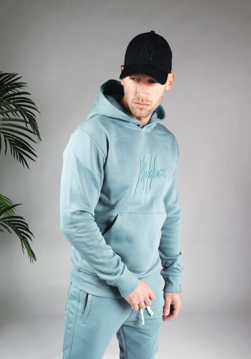 Vooraanzicht van model gekleed in een blauwe hoodie met het geborduurde logo van Malelions in het blauw in combinatie met de blauwe trainingsbroek en een zwarte pet. Het model heeft zijn armen losjes langs zich en kijkt schuin naar de grond.