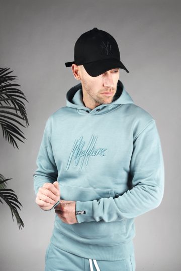 Vooraanzicht van model gekleed in een blauwe hoodie met het geborduurde logo van Malelions in het blauw in combinatie met de blauwe trainingsbroek en een zwarte pet. Het model heeft zijn handen voor zich en kijkt langs zijn schouder naar links.