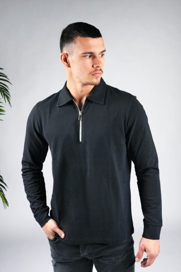Vooraanzicht van model gekleed in een zwarte longsleeve polo trui met halve rits. Het model heeft een hand in zijn zak en kijkt schuin naar links.