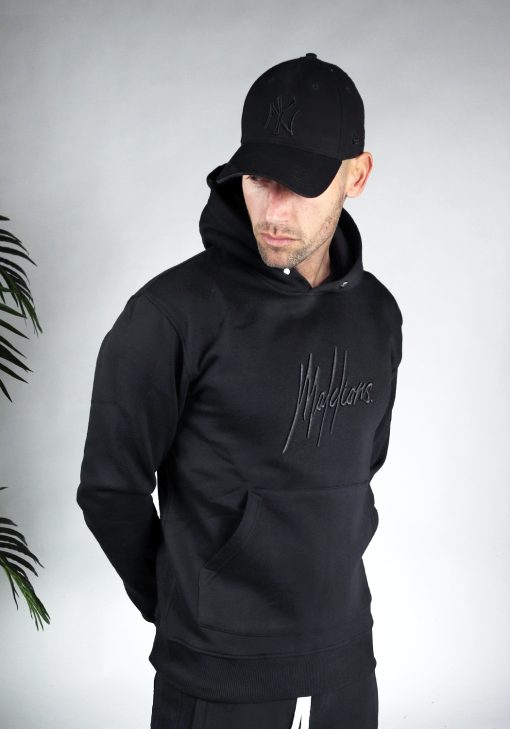 Vooraanzicht van model gekleed in zwarte hoodie met een zwart geborduurd logo van Malelions op de borst. Het model heeft zijn handen achter zijn rug en kijkt schuin naar de grond.