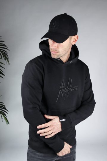 Vooraanzicht van model gekleed in zwarte hoodie met een zwart geborduurd logo van Malelions op de borst. Het model pakt zijn eigen arm vast en kijkt naar de grond.