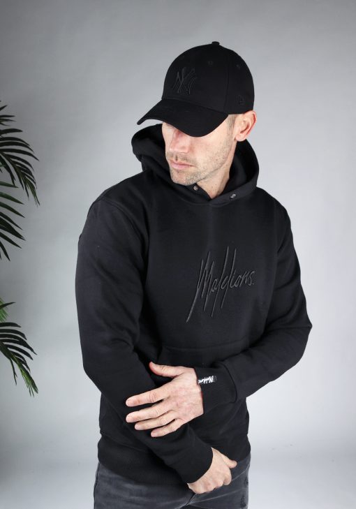 Vooraanzicht van model gekleed in zwarte hoodie met een zwart geborduurd logo van Malelions op de borst. Het model pakt zijn eigen arm vast en kijkt naar de grond.