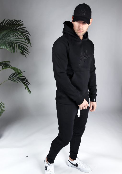 Zijaanzicht van model gekleed in een zwarte hoodie met het geborduurde logo van Malelions in het zwart in combinatie met de blauwe trainingsbroek en een zwarte pet. Het model heeft zijn armen losjes langs zich en kijkt schuin naar de grond.