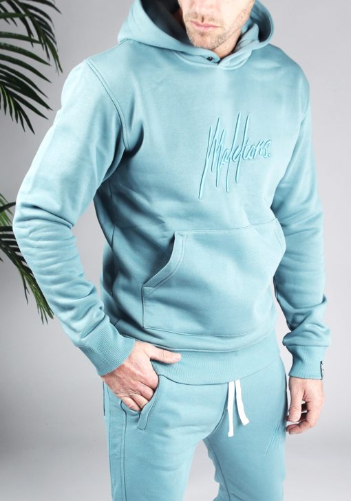 Vooraanzicht van model gekleed in blauwe joggerset met het blauw geborduurde Malelions logo op de borst en het bovenbeen.