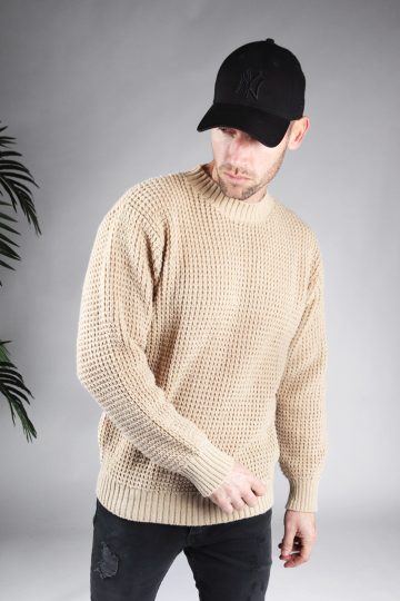 Schuin vooraanzicht van model gekleed in een beige warme knit sweater met een oversized fit in combinatie met een zwarte broek en een zwarte pet. Het model heeft zijn handen losjes naast zich en kijkt schuin naar de grond.