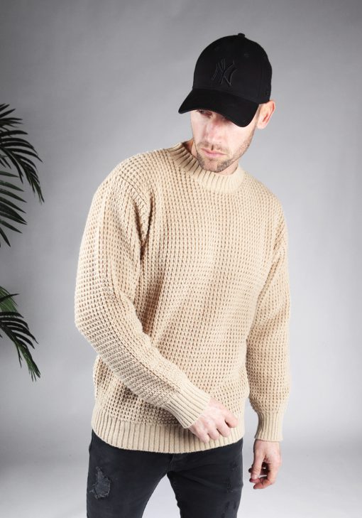 Schuin vooraanzicht van model gekleed in een beige warme knit sweater met een oversized fit in combinatie met een zwarte broek en een zwarte pet. Het model heeft zijn handen losjes naast zich en kijkt schuin naar de grond.