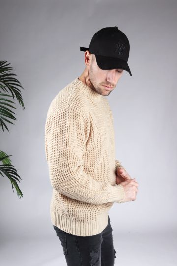 Rechter zijaanzicht van model gekleed in een beige warme knit sweater met een oversized fit in combinatie met een zwarte broek en een zwarte pet. Het model heeft zijn handen voor zich en kijkt schuin naar de grond.