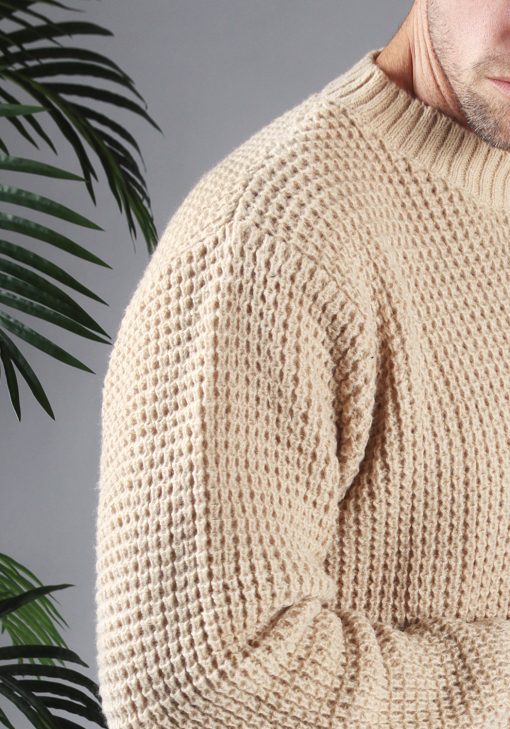 Close up van model gekleed in een beige knit sweater met een oversized fit, waarop het gehaakte patroon van de trui goed zichtbaar is.