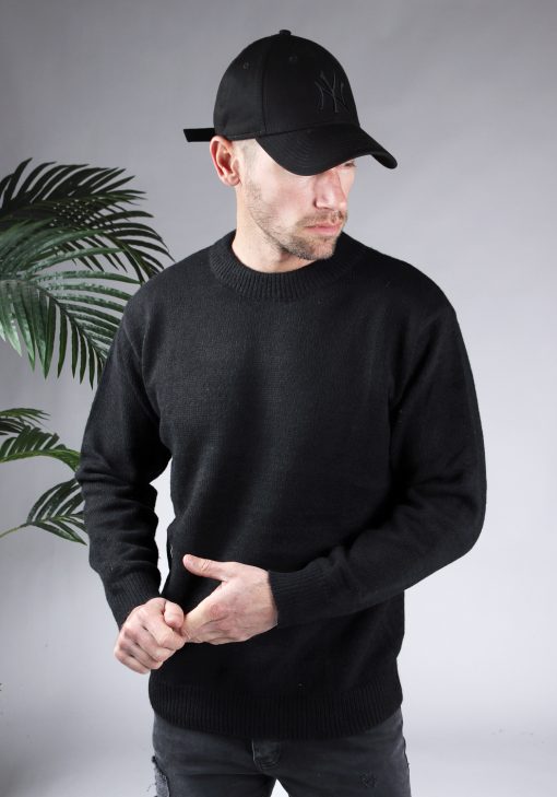 Vooraanzicht van model gekleed in een zwarte oversized knit sweater in combinatie met een zwarte broek en een zwarte pet. Het model heeft zijn handen voor zich en kijkt schuin naar de grond.