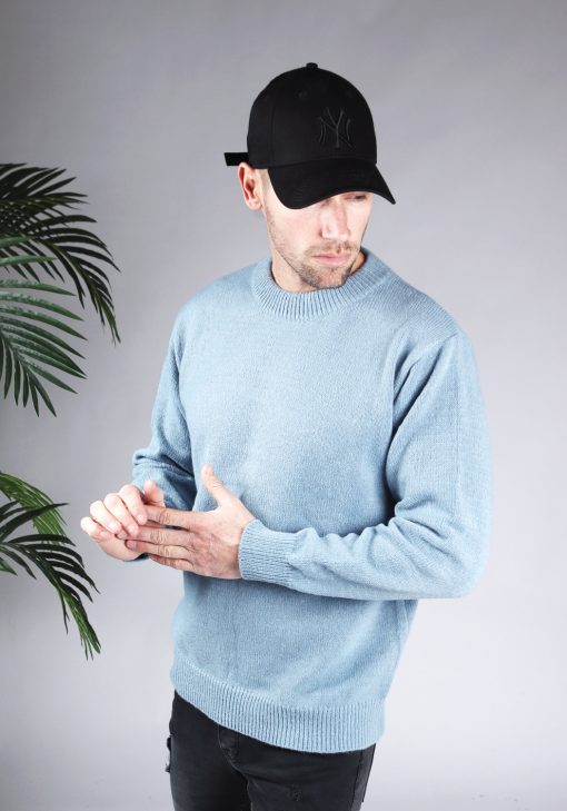 Vooraanzicht van model gekleed in een lichtblauwe oversized knit sweater in combinatie met een zwarte broek en een zwarte pet. Het model heeft zijn handen voor zich en kijkt schuin naar de grond.