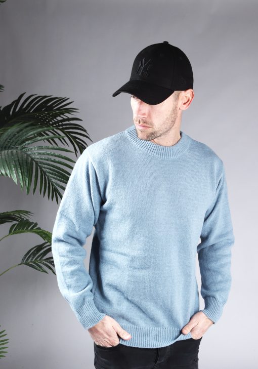 Vooraanzicht van model gekleed in een lichtblauwe oversized knit sweater in combinatie met een zwarte broek en een zwarte pet. Het model heeft zijn handen in zijn voorzakken en kijkt langs zijn schouder naar de grond.