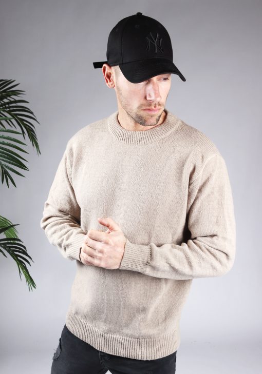 Vooraanzicht van model gekleed in een lichtbruine knit sweater in combinatie met een zwarte broek en een zwarte pet. Het model heeft zijn handen voor zich en kijkt schuin naar de grond.
