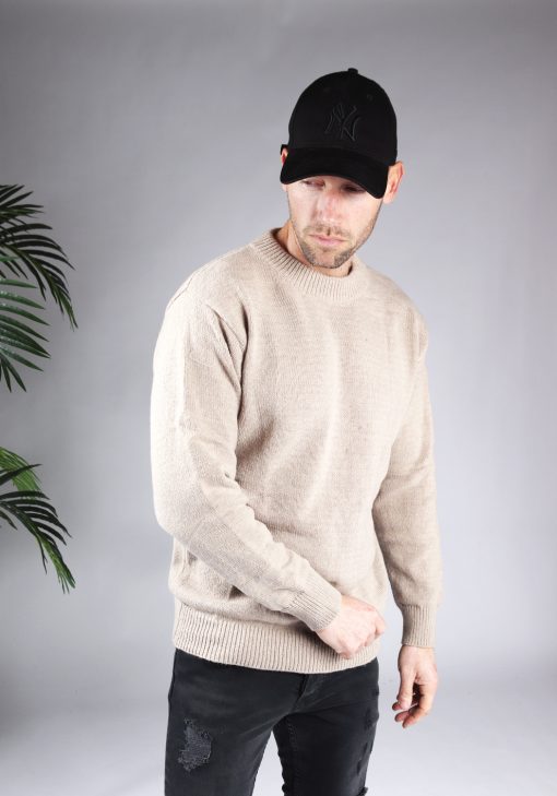 Zijaanzicht van model gekleed in een lichtbruine knit sweater in combinatie met een zwarte broek en een zwarte pet. Het model heeft zijn handen losjes naast zich en kijkt schuin naar de grond.