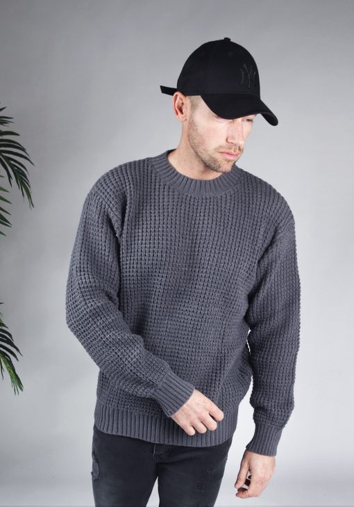 Vooraanzicht van model gekleed in een grijze warme knit sweater met een oversized fit in combinatie met een zwarte broek en een zwarte pet. Het model heeft zijn handen losjes naast zich en kijkt schuin naar de grond.