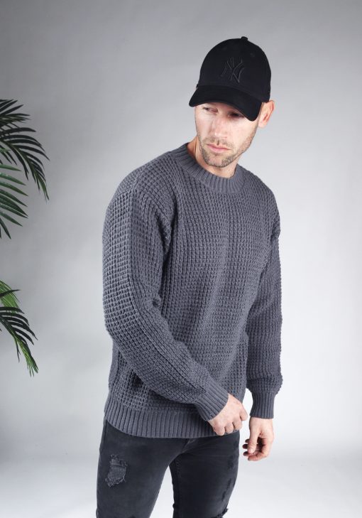 Zijaanzicht van model gekleed in een grijze knit sweater in combinatie met een zwarte broek en een zwarte pet. Het model heeft zijn handen losjes naast zich en kijkt schuin naar de grond.