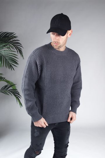Rechter zijaanzicht van model gekleed in een grijze knit sweater in combinatie met een zwarte broek en een zwarte pet. Het model heeft zijn handen aan de onderkant van de trui en kijkt schuin naar de grond.