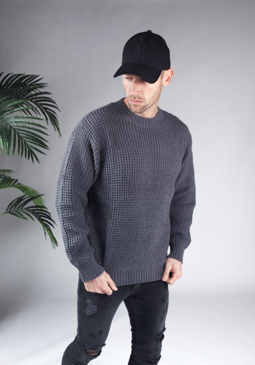 Rechter zijaanzicht van model gekleed in een grijze knit sweater in combinatie met een zwarte broek en een zwarte pet. Het model heeft zijn handen aan de onderkant van de trui en kijkt schuin naar de grond.