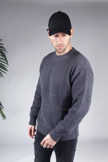 Linker zijaanzicht van model gekleed in een grijze knit sweater in combinatie met een zwarte broek en een zwarte pet. Het model heeft zijn handen losjes naast zich en kijkt schuin naar de grond.