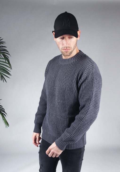 Linker zijaanzicht van model gekleed in een grijze knit sweater in combinatie met een zwarte broek en een zwarte pet. Het model heeft zijn handen losjes naast zich en kijkt schuin naar de grond.