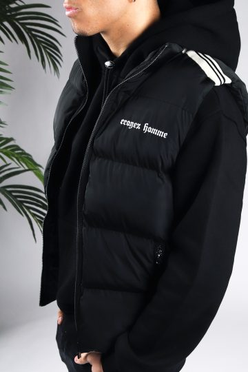 Close up van model gekleed in een zwarte bodywarmer met witte details in combinatie met een zwarte hoodie. De bodywarmer heeft witte tekst op de linkerborst en details op de schouder. Het model heeft zijn handen in zijn zakken.