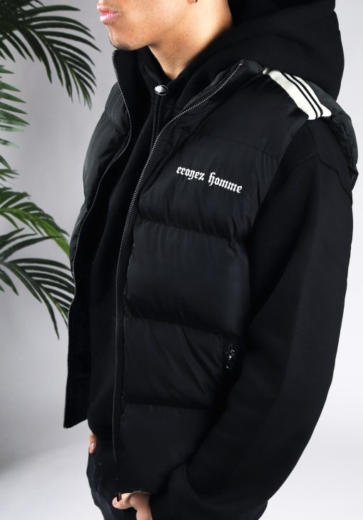 Close up van model gekleed in een zwarte bodywarmer met witte details in combinatie met een zwarte hoodie. De bodywarmer heeft witte tekst op de linkerborst en details op de schouder. Het model heeft zijn handen in zijn zakken.