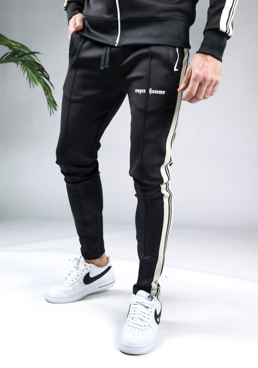 Close up van de broek van een zwarte Croyez tracksuit gecombineerd met zwart-witte Nike sneakers. Het pak heeft witte lijnen langs de armen en benen en witte tekst op de bovenbeen. Het model heeft een hand in zijn zak.