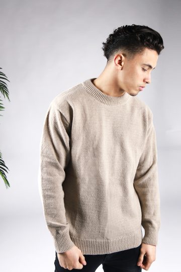 Vooraanzicht van model gekleed in een lichtbruine knit sweater in combinatie met een zwarte broek. Het model heeft zijn handen langs zijn lichaam en kijkt links naar de grond.