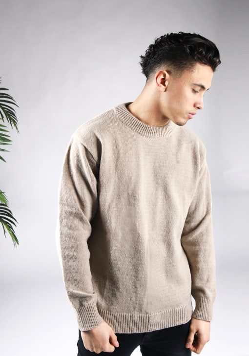 Vooraanzicht van model gekleed in een lichtbruine knit sweater in combinatie met een zwarte broek. Het model heeft zijn handen langs zijn lichaam en kijkt links naar de grond.