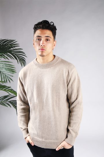 Vooraanzicht van model gekleed in een lichtbruine knit sweater met een oversized fit in combinatie met een zwarte broek. Het model heeft zijn handen in zijn voorzakken en kijkt recht in de camera.